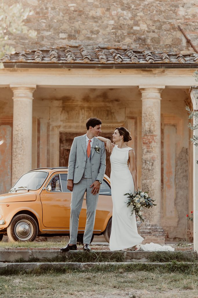A bride in a white gown and bouquet stands arm-in-arm with her groom in a light gray suit and red tie. Captured by a Wedding Photographer Florence, they pose before an old building with columns and an orange vintage car.
