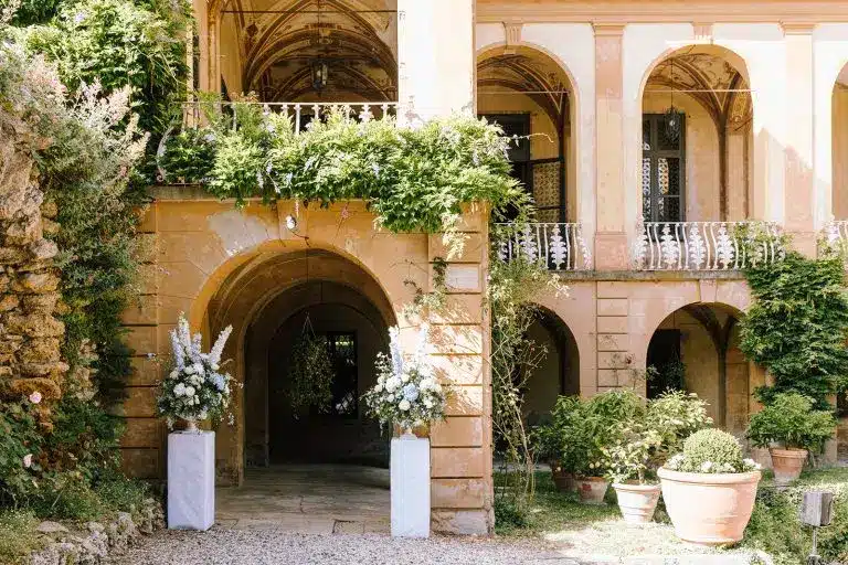 Arched entrance of the elegant villa di striano with climbing greenery, potted plants, and two tall flower arrangements on white pedestals, framed by warm yellow walls and a balcony above.