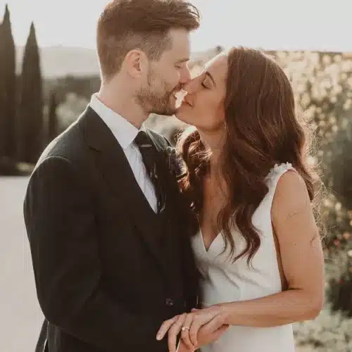 A bride and groom share a kiss outdoors The groom wears a black suit and tie, while the bride wears a sleeveless white dress. They embrace on a sunlit path with greenery in the background.