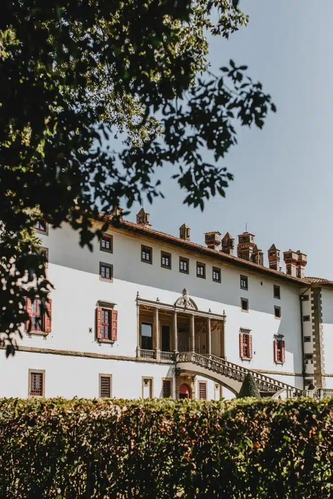 A grand, historic building—Tenuta di Artimino—with white walls, red shutters, and ornate chimneys, is partially framed by tree branches and greenery under a clear sky.
