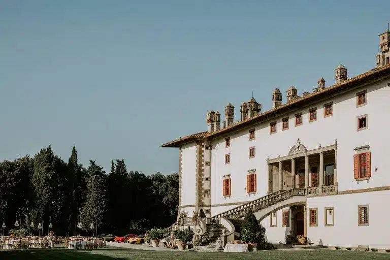 A large historic villa, Tenuta di Artimino, with cream-colored walls, red shutters, and ornate details stands among green trees and a manicured lawn under a blue sky. Outdoor tables and chairs are set up on the grass.