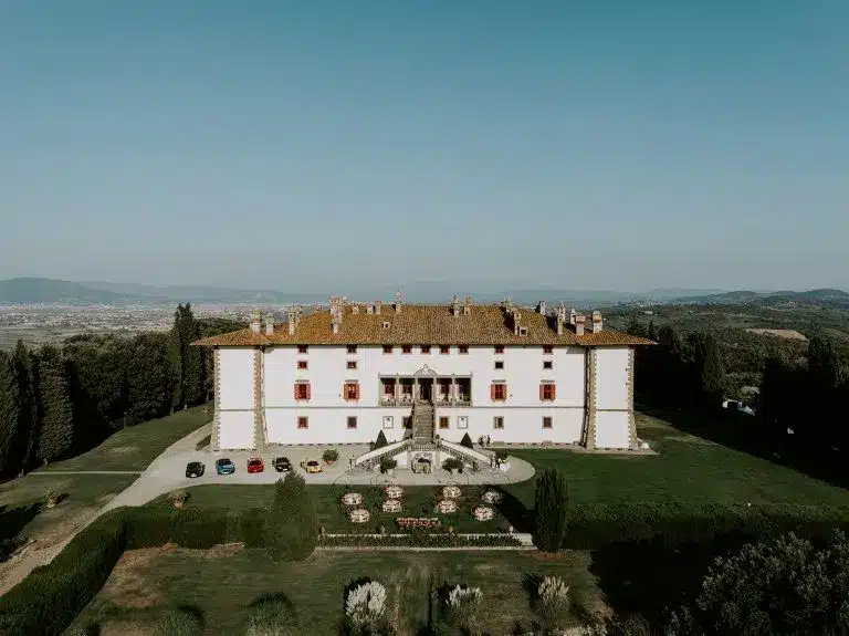 Aerial view of Tenuta di Artimino, a large, historic villa with a red-tiled roof, surrounded by green lawns and gardens, cars parked near the entrance, and rolling hills visible in the distance under a clear blue sky.