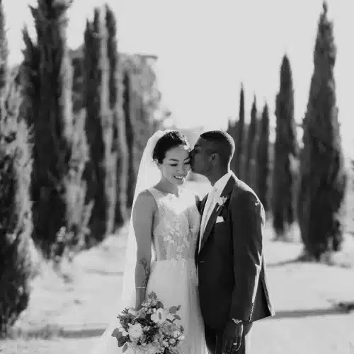 A bride and groom stand outdoors on a tree-lined path. The groom kisses the bride’s temple while she smiles, holding her bouquet—a perfect example of Tips for Wedding Photography Poses: How to Look Natural. They both look joyful in this black and white image.