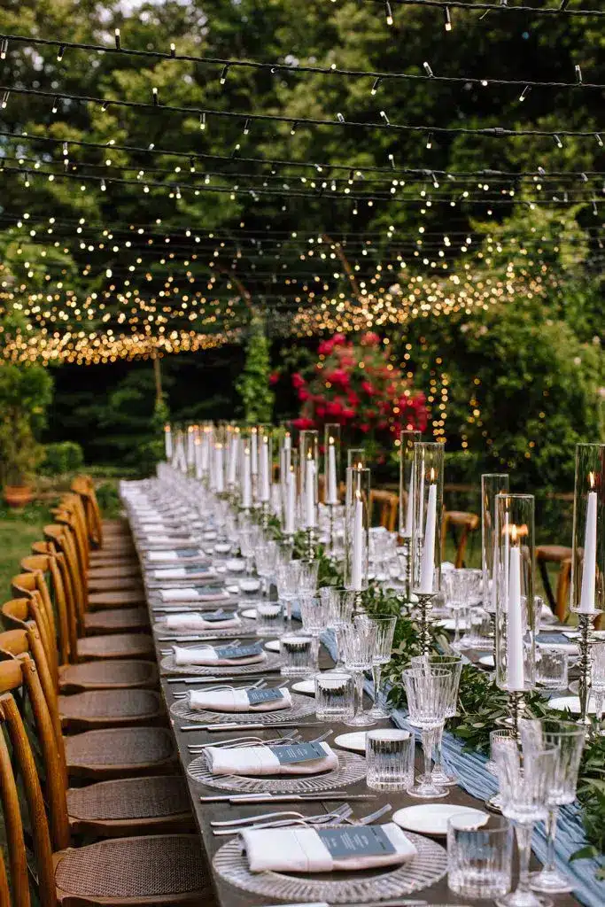A long outdoor dining table is elegantly set with candles, glassware, and plates. Overhead, strings of warm fairy lights glow at Villa di Striano, with lush greenery and trees creating a festive, romantic atmosphere.
