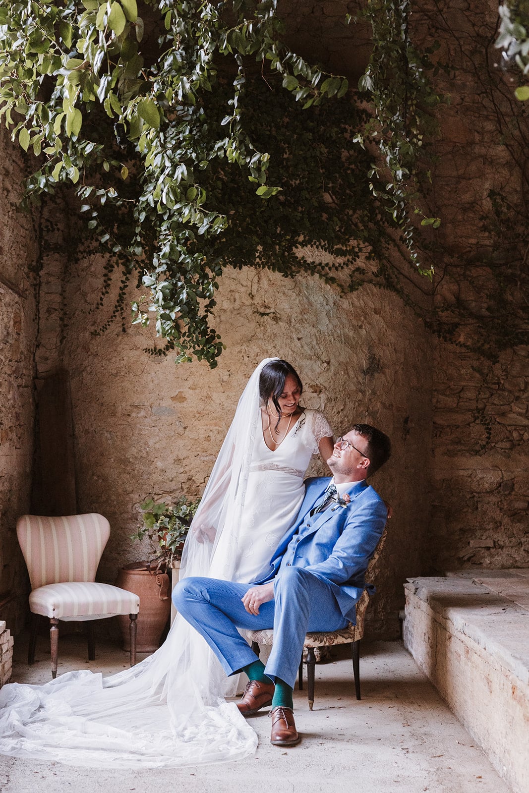 A bride in a white dress and veil stands beside a seated groom in a blue suit. They gaze at each other, smiling, in the rustic stone room of Villa di Striano, adorned with hanging greenery and a striped chair nearby.