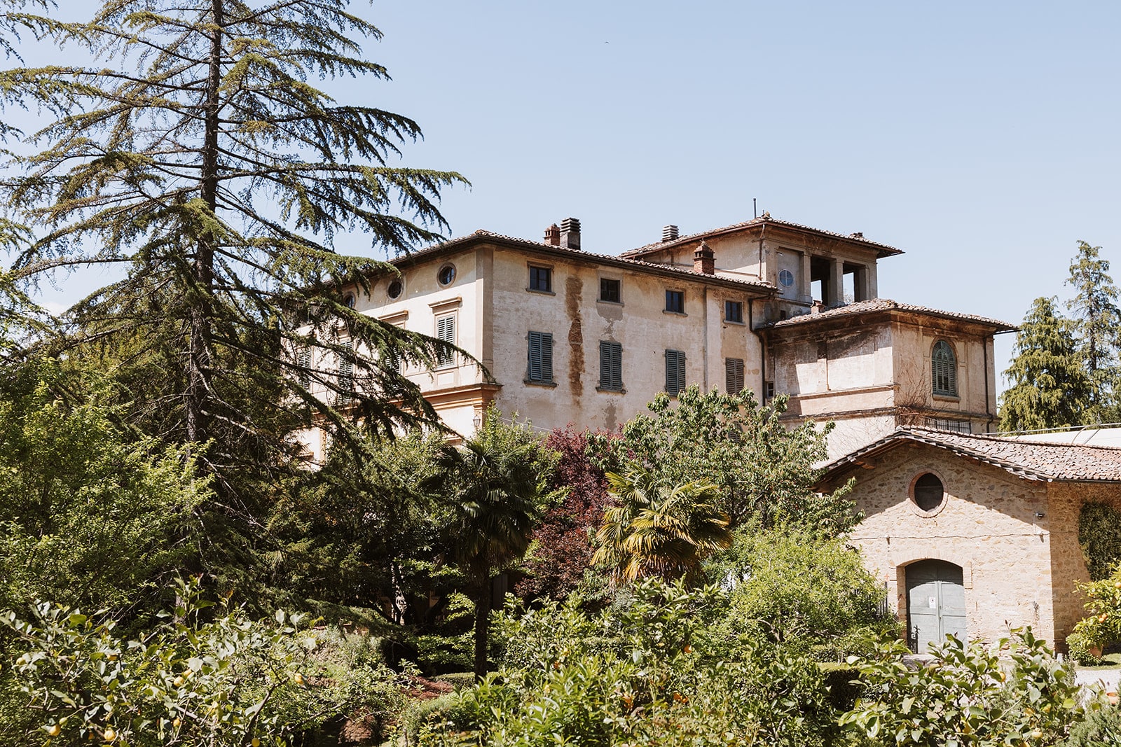 Large, rustic Italian villa di striano with tan stucco walls and green shutters, surrounded by lush trees, plants, and greenery under a clear blue sky.