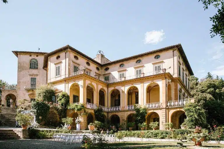 A historic, yellow-walled villa di striano with arched balconies and lush greenery, set under a clear sky. Several white chairs are arranged in rows on the lawn in front, suggesting an outdoor event or ceremony.