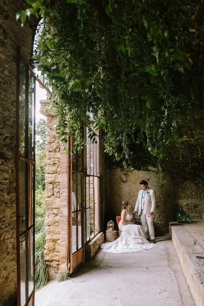 A bride in a white dress sits beside a groom in a light-colored suit inside Villa di Striano, a rustic stone building with tall windows and lush greenery overhead. Natural light streams in, creating a serene and romantic atmosphere.