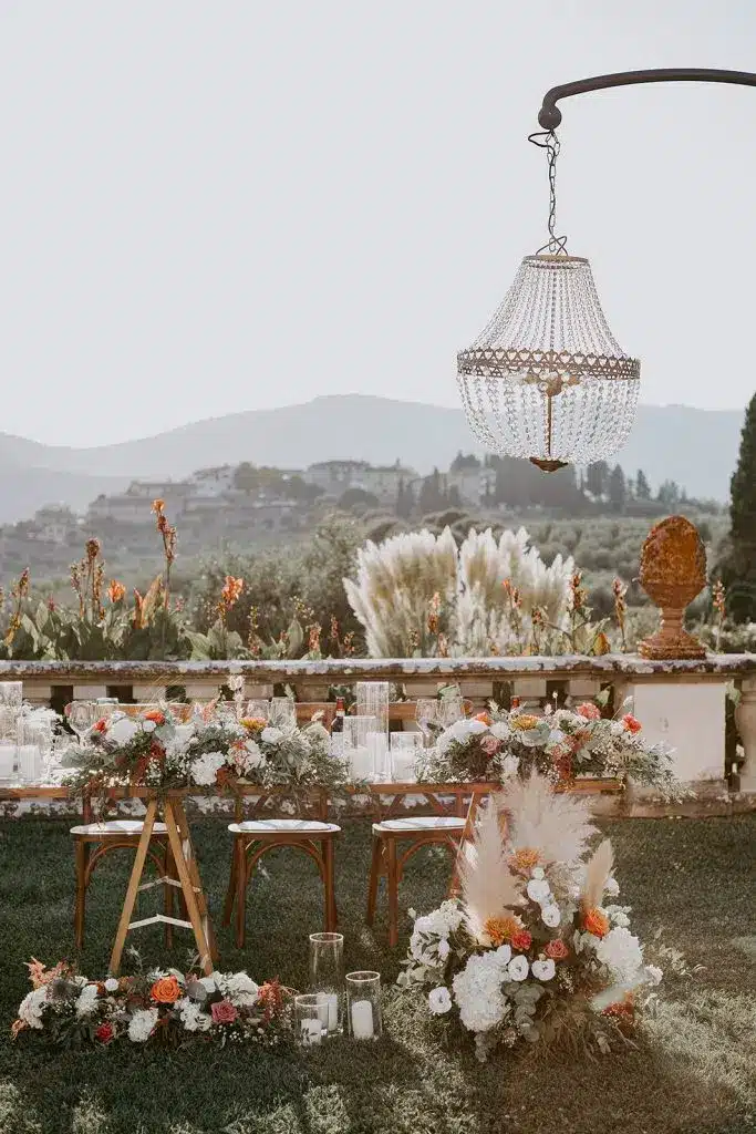 An outdoor wedding table at Tenuta di Artimino is set with floral arrangements, candles, and wooden chairs beneath a crystal chandelier, with stunning mountains and greenery gracing the background.