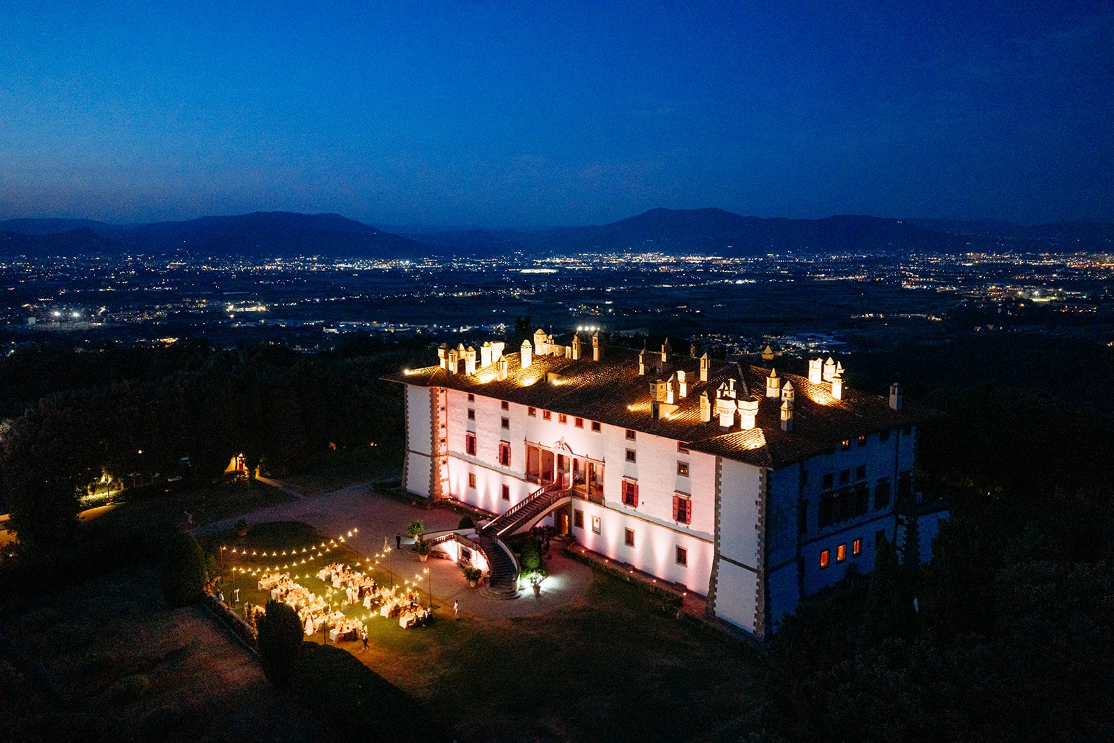 A large, illuminated villa at Tenuta di Artimino hosts an outdoor evening event with tables set up on the lawn; city lights and mountains are visible in the distance under a deep blue sky.