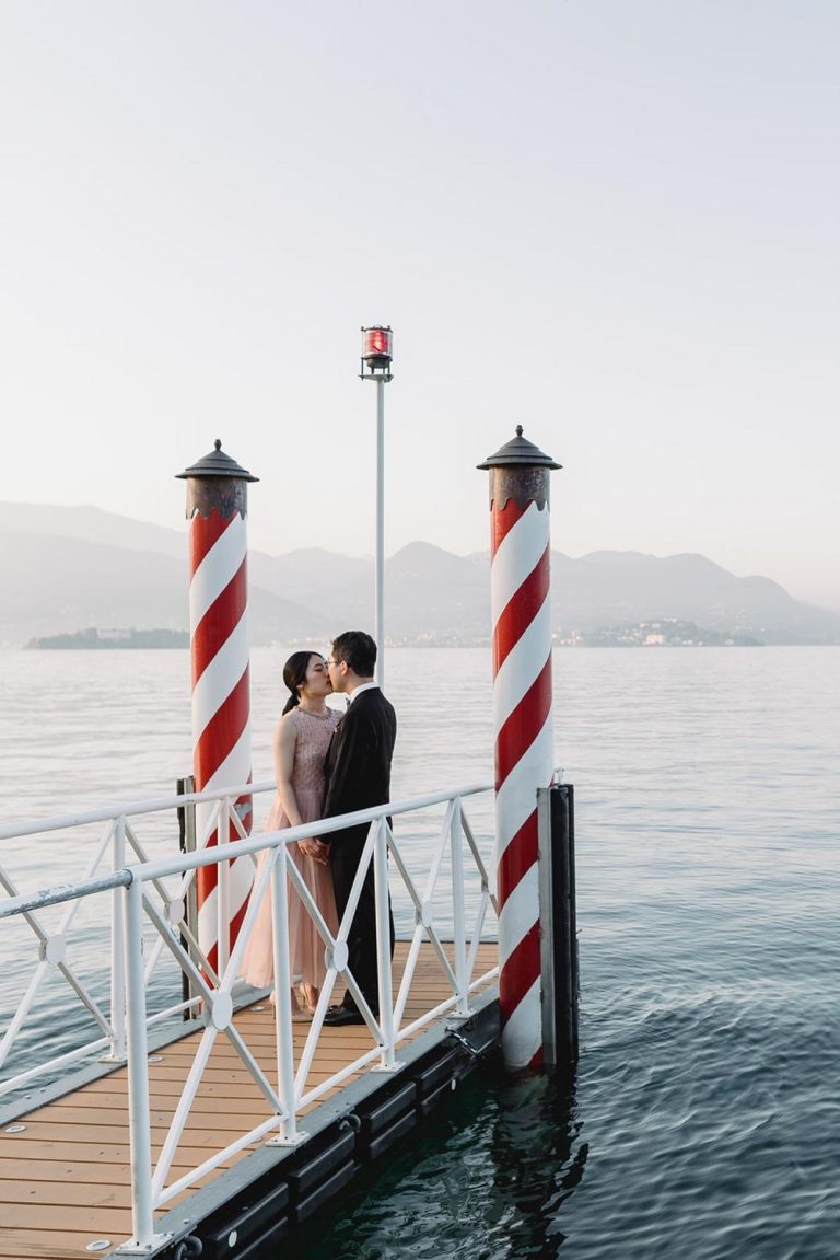 A couple stands close together on a dock over calm water, framed by two red-and-white striped poles, surrounded by mountains and a hazy sky—the-people-you-love-in-the-same-place, sharing a peaceful moment.