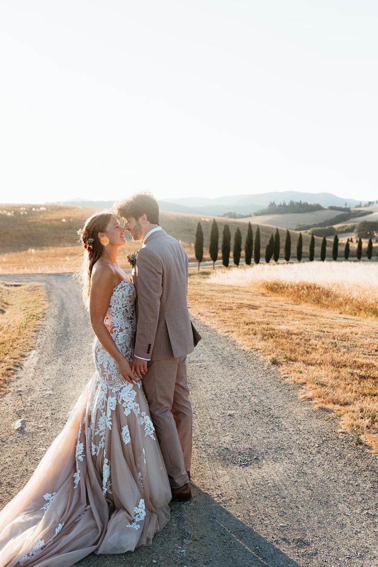 A bride and groom stand on a dirt road, holding hands and kissing beneath a bright sky—an inspiring scene for those wondering how to choose a destination wedding photographer in Italy to capture your own golden field romance.